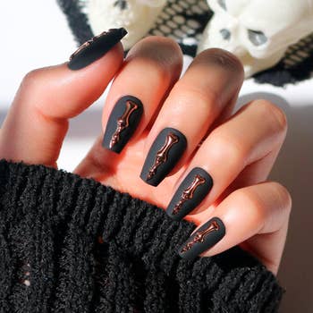 A manus with matte achromatic coffin-shaped nails featuring shiny metallic skeleton designs, against a textured cloth background