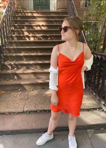 Reviewer wearing short orange silk dress with cowl neckline, layered gold necklaces, and a white long sleep button down in front of stairs