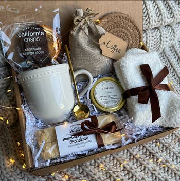 Product open with package of biscotti, package of chocolate covered orange slices, white mug, golden spoon, bag of coffee, gold candle tin, white fuzzy socks, and a woven coaster