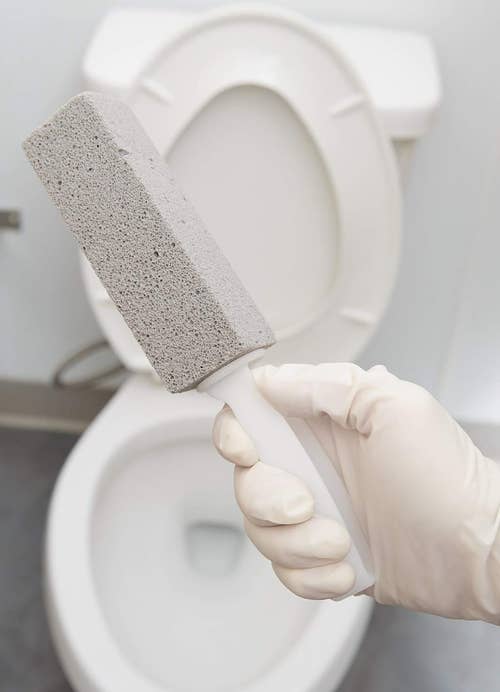 Hand in a glove holding a pumice cleaning stone near an open toilet bowl
