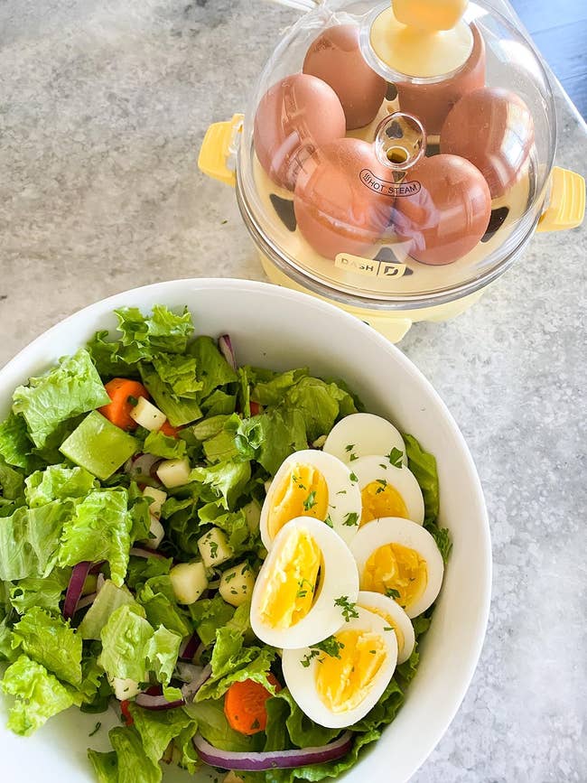 Salad with lettuce, cucumbers, and carrots successful a bowl, topped with sliced boiled eggs; ovum steamer with boiled eggs successful the background