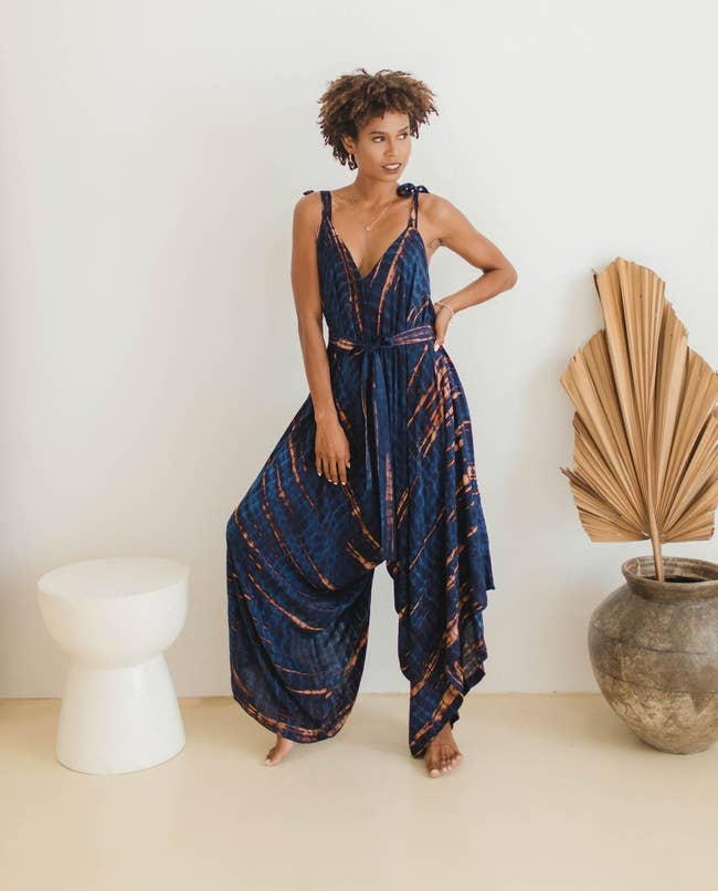 Person successful a stylish, flowing jumpsuit stands by a modern stool and vase with dried leaves