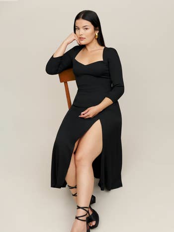 a model sitting while wearing a black midi dress and sandal heels