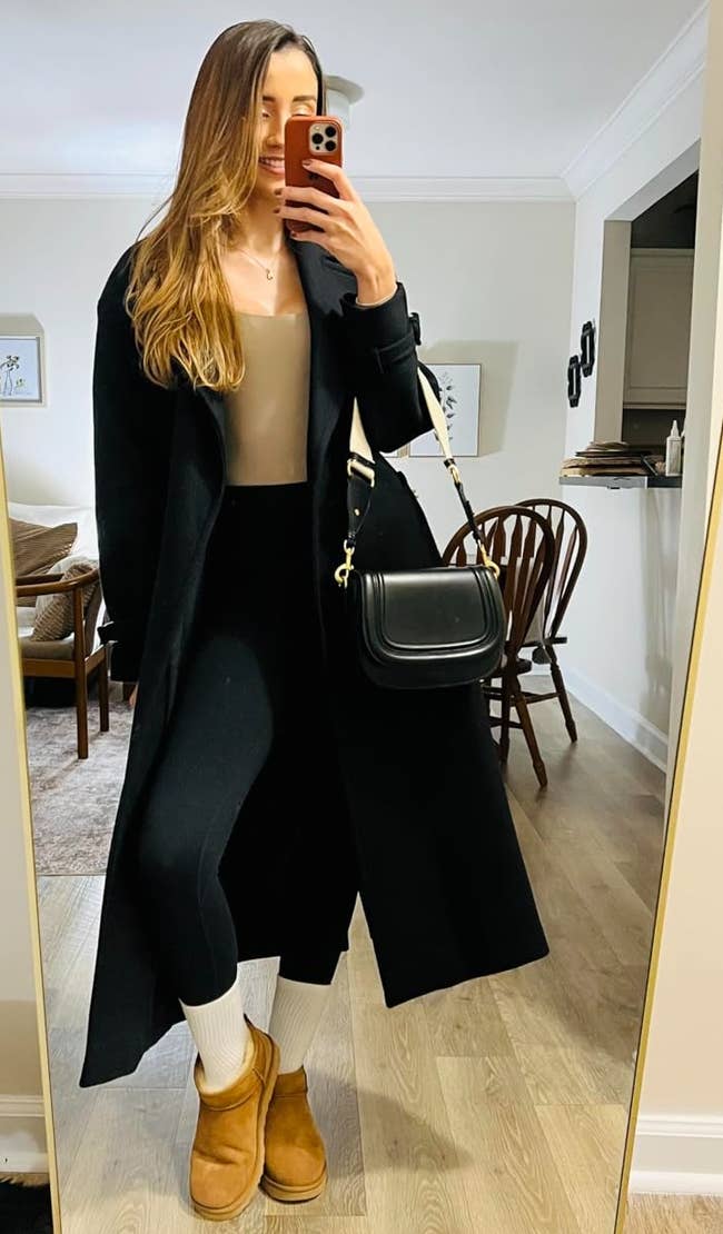 Person taking a reflector selfie, wearing a agelong coat, leggings, boots, and carrying a handbag