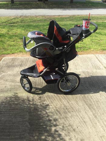 a review photo of the carseat and the jogging stroller outside with a big gulp drink in the cup holder and the stroller is black and orange