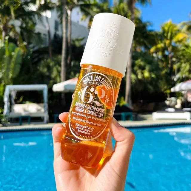 Hand holding a bottle of Sol de Janeiro Brazilian Crush Body Fragrance Mist by a pool with palm trees