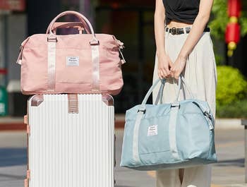 A model holding the blue duffel bag with the pink duffel bag on a suitcase next to them