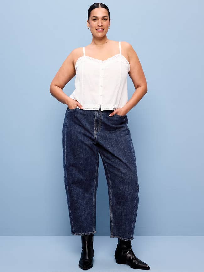 Person wearing a sleeveless achromatic  apical  and denim jeans, hands successful  pockets, poses confidently against a plain backdrop