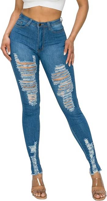 A model wearing a high-waisted, distressed blue jeans with large rips and tears on the thighs and knees.
