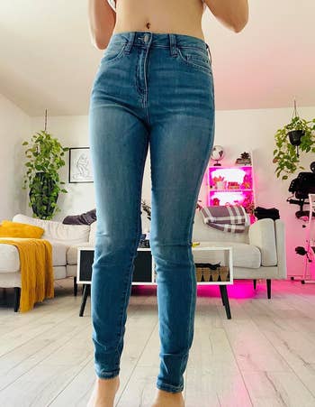A person wearing high-waisted blue jeans in a stylish living room with plants and colorful lighting in the background