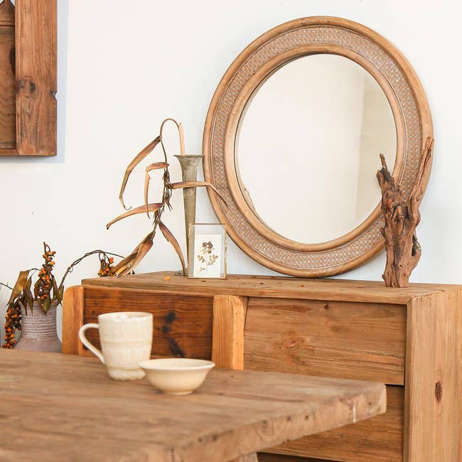 A rustic wooden sideboard with a circular mirror, decorative vase, and cup staged in a cozy interior for home decor inspiration