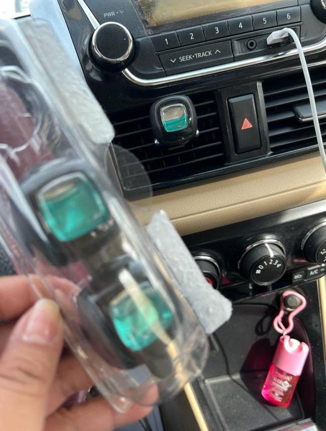 Reviewer holding three pack of small square clip-on scents with one of them attached to the AC vent 