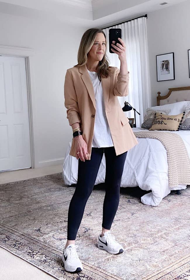 Person taking a mirror selfie, wearing a casual blazer, white top, dark leggings, and sneakers, in a stylishly decorated bedroom
