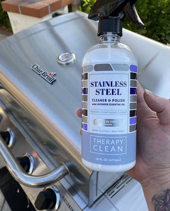 reviewer holding the bottle of stainless steel cleaning spray next to a stainless steel grill