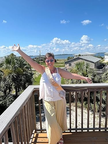 reviewer on a balcony with arms outstretched, wearing a sash, wide-legged pants, and sunglasses, with palm trees and a coastal view in the background