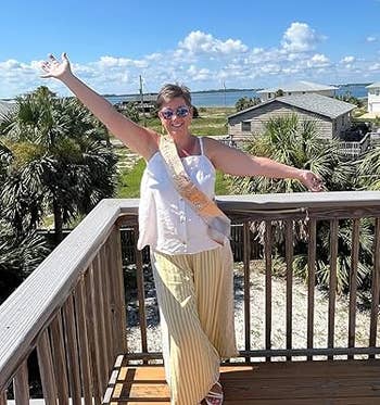 reviewer on a balcony with arms outstretched, wearing a sash, wide-legged pants, and sunglasses, with palm trees and a coastal view in the background