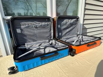 Two unfastened  hard-shell suitcases, 1  bluish  and 1  orange, displayed connected  a table, showcasing spacious interiors and compartments