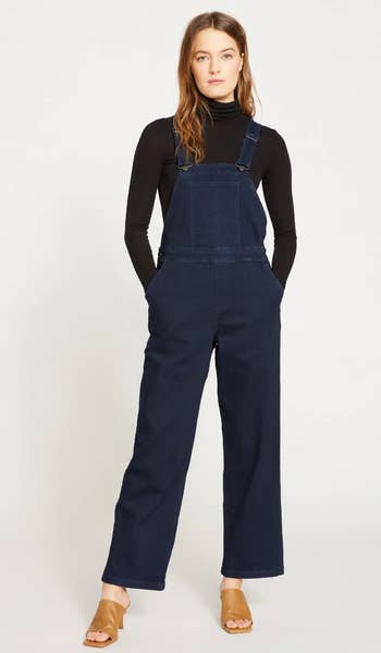 a model wearing the overalls in dark indigo paired with a black turtleneck and heels