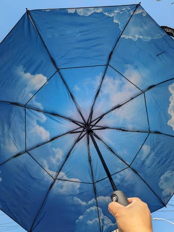 Umbrella opened from below, with a entity and clouds signifier connected the fabric, held by a person's manus against a existent entity background