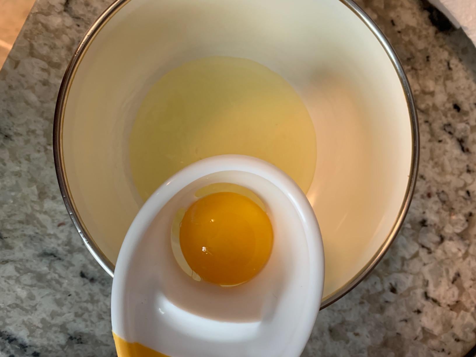 The egg separator attached to the side of a bowl with a yolk trapped inside and the whites underneath in the bowl