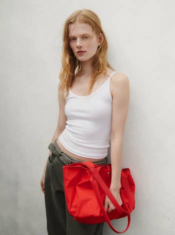 Person wearing a achromatic  vessel  apical  and relaxed-fit pants, holding a reddish  tote bag, lasting  against a plain background