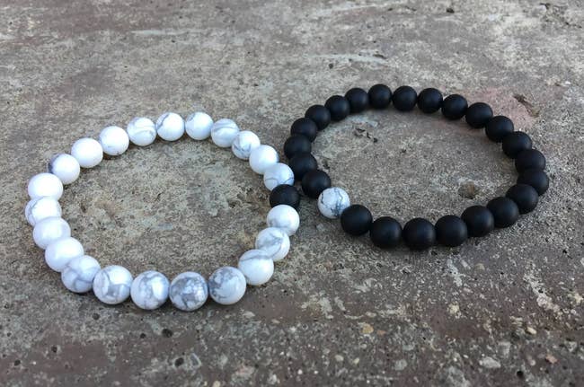 White marble patterned beaded bracelet with single black bead next to a black beaded bracelet with a single white marble bead on top of rock