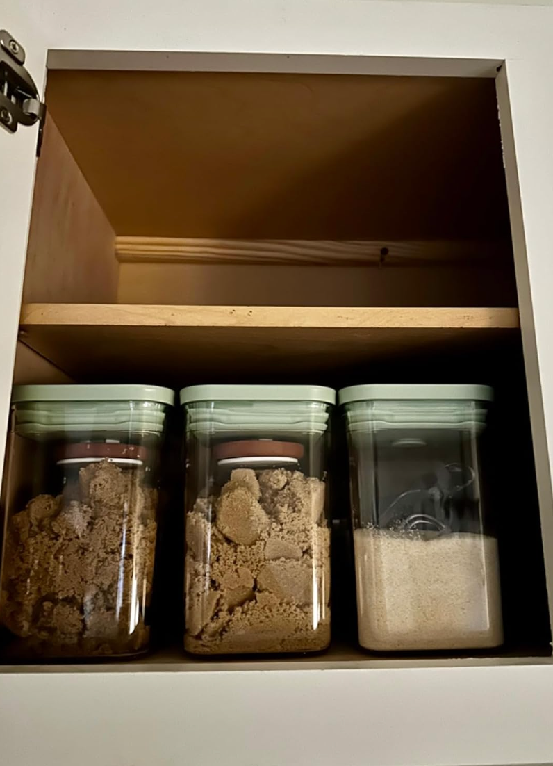 Three kitchen containers in a cabinet, two filled with brown sugar and one with white sugar, topped with round brown items