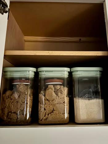 Three kitchen containers in a cabinet, two filled with brown sugar and one with white sugar, topped with round brown items