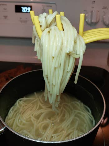 Spaghetti noodles being lifted from a pot of boiling water with a yellow kitchen tool. Stove clock shows 1:09