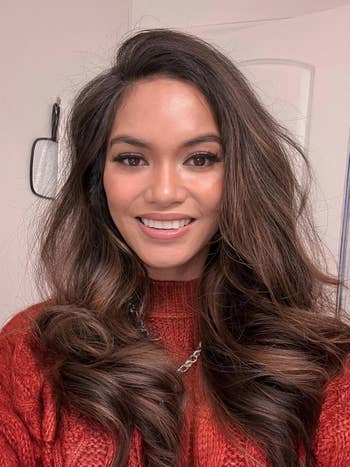 Peninjau tersenyum, mengenakan sweater bertekstur dan kalung rantai. Rambut panjang dan bergaya. Gambar untuk Artikel Belanja