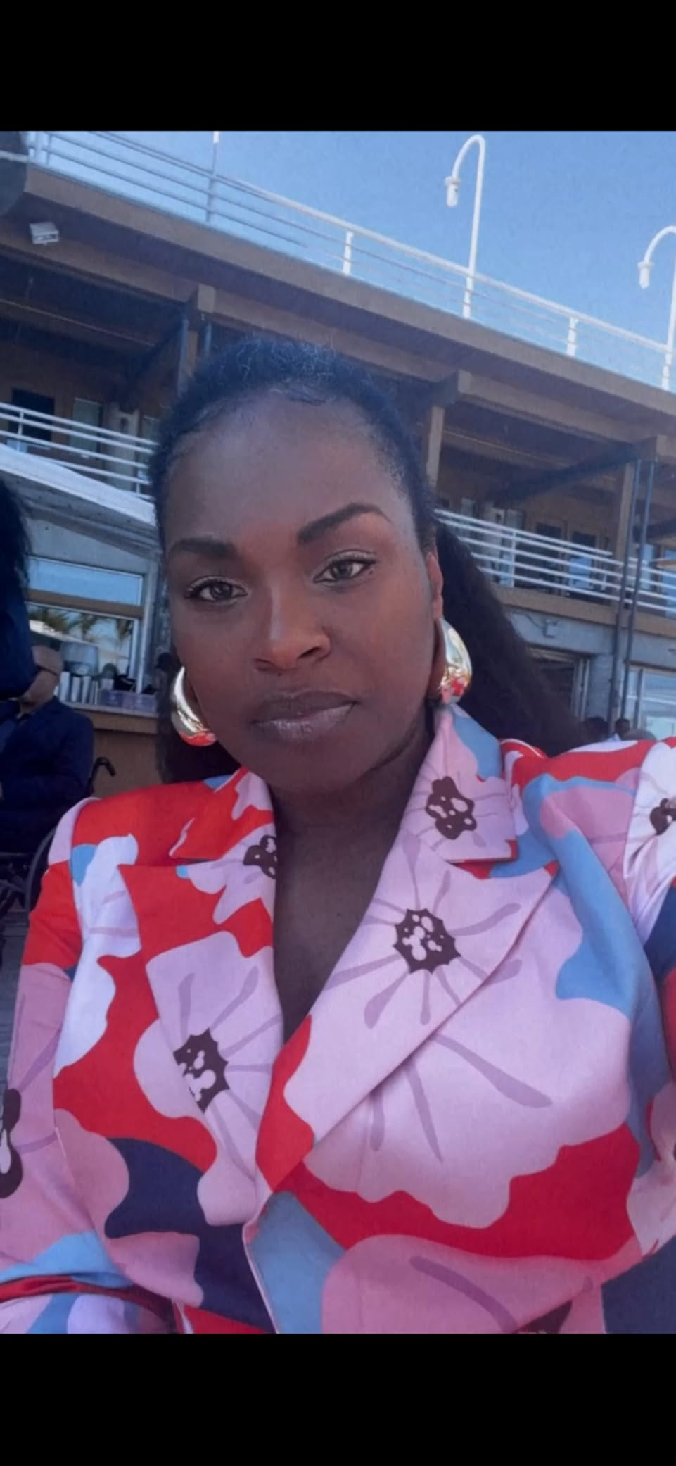 Person wearing a floral-patterned blouse with ample  hoop earrings, posing outdoors