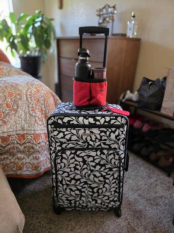 Floral-patterned suitcase with two travel mugs in a red holder sits on a carpeted bedroom floor near a bed and dresser