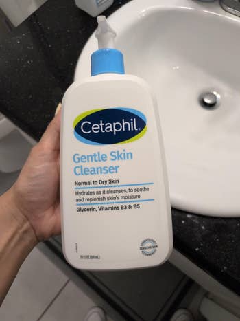 A manus  holds a vessel  of Cetaphil Gentle Skin Cleanser by a bath  sink. The statement   highlights benefits similar  hydration and skincare with vitamins B3 and B5