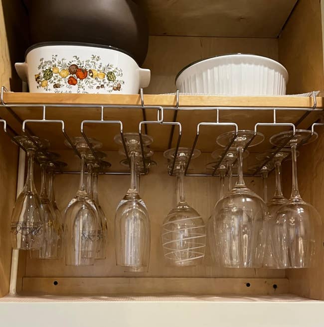 Kitchen cabinet with hanging stemware rack, holding various wine glasses. Above are stacked ceramic dishes, including a floral-patterned casserole dish