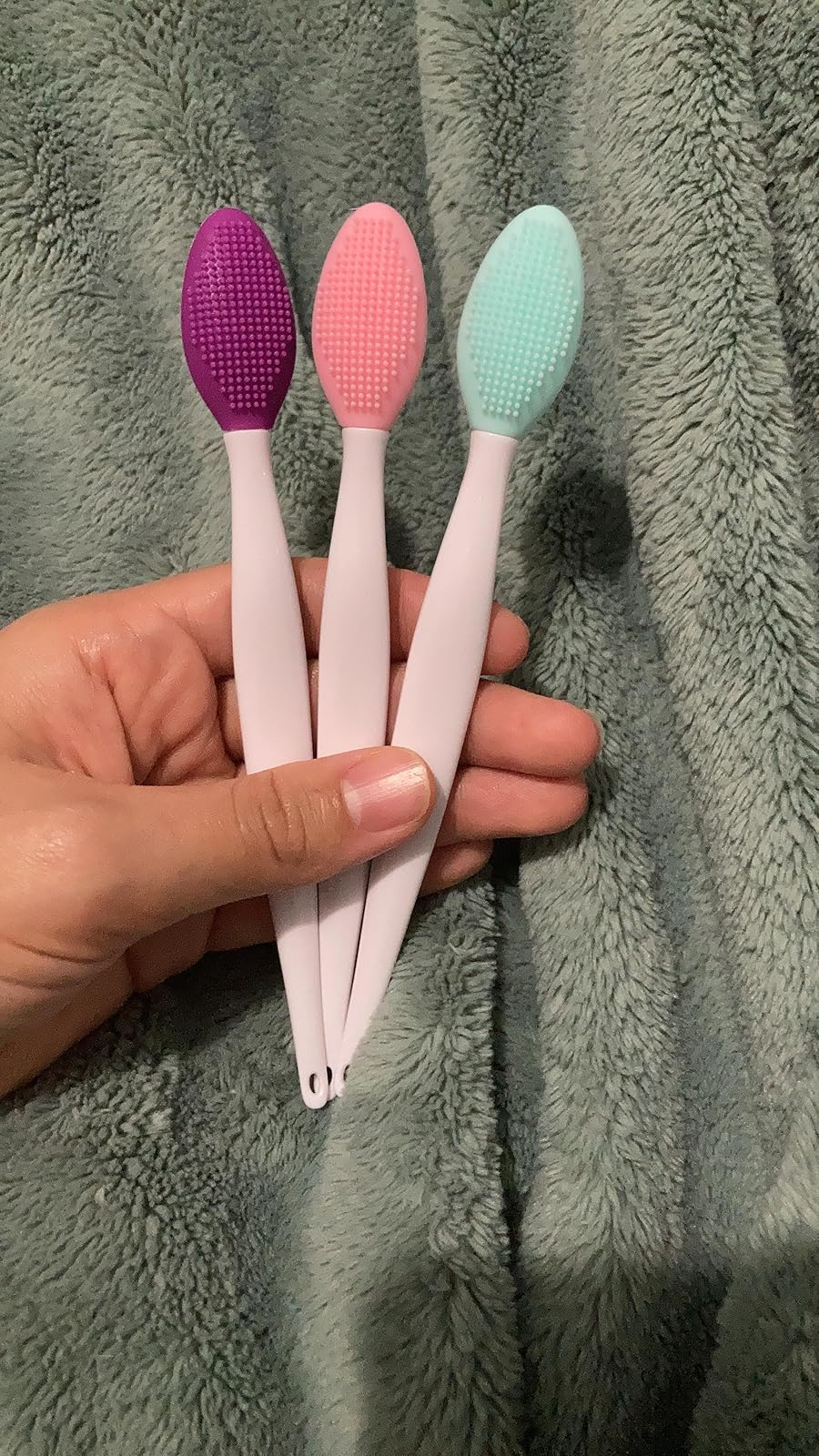 Three silicone exfoliating brushes in purple, pink, and teal held in a hand, placed on a textured fabric background