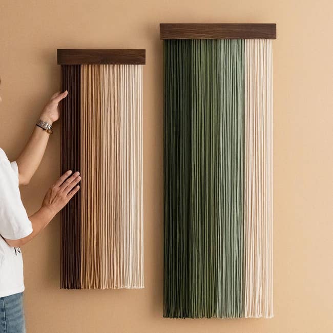 Person adjusting 2 vertical partition hangings made of strands, 1 brownish gradient and the different greenish gradient, connected a tan wall