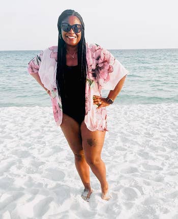 Person smiling astatine  the beach, wearing a floral cover-up implicit    a achromatic  swimsuit, sunglasses, and a smartwatch
