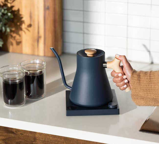 a hand reaching for the blue and maple wood electric kettle on a countertop