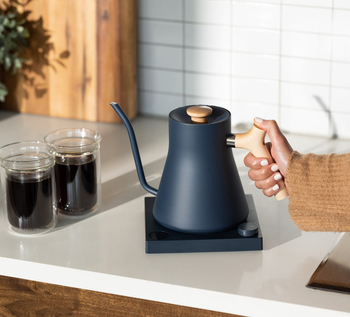 a hand reaching for the blue and maple wood electric kettle on a countertop