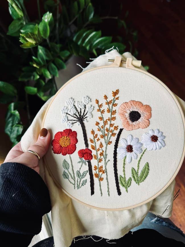 Person holding an embroidery hoop with elaborate floral designs, including daisies and poppies, adjacent to a leafy plant