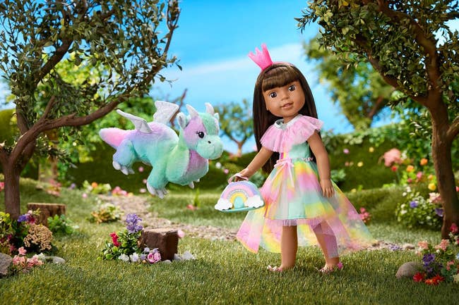 A doll wearing a tulle formal  and crown stands successful  a plot  with a plush dragon, holding a rainbow-themed purse