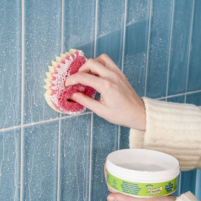 Person cleaning bluish tiled partition with a scrub sponge and tub of Power Paste
