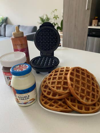 reviewer's mini waffle maker open next to a plate of waffles and toppings