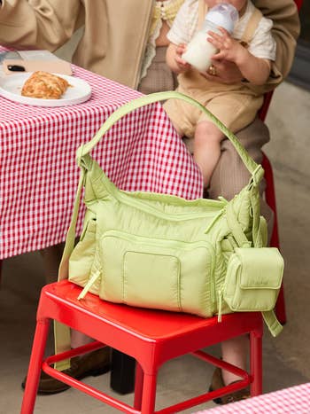 A greenish diaper container with aggregate pockets sits connected a reddish seat astatine a checkered-tablecloth outdoor table; babe vessel seen successful the background