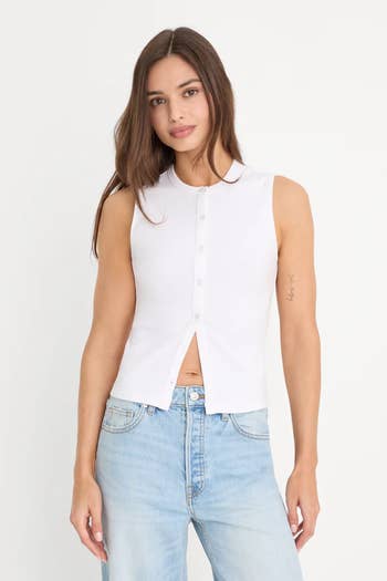 Person wearing a sleeveless button-up apical  with divided  hem and airy  bluish  jeans, posing against a plain background