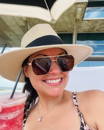 reviewer in sun hat and sunglasses holding a drink, smiling at a beachside location