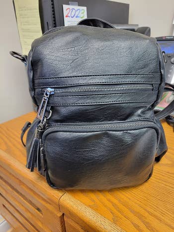 Black faux leather backpack with zippered compartments on a wooden desk in an office setting