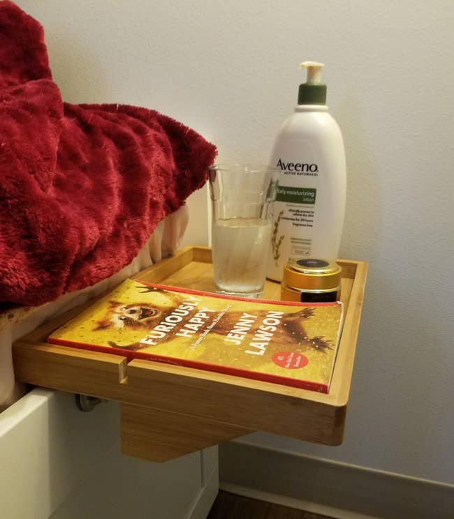A light wood shelf attached to a bed frame holding a book, water, and beauty supplies 