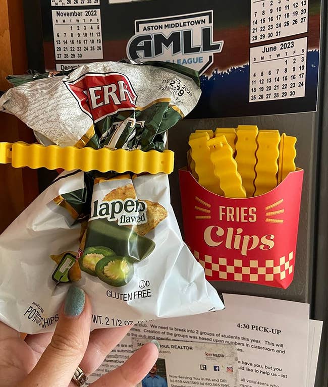 image of reviewer holding up an open bag of chips with a french fry clip on it next to the container of french fry clips stuck on their fridge