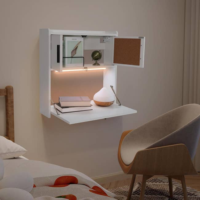 Wall-mounted white desk with lamp, globe, book, and corkboard; situated above a bed beside a chair, suitable for small space home offices